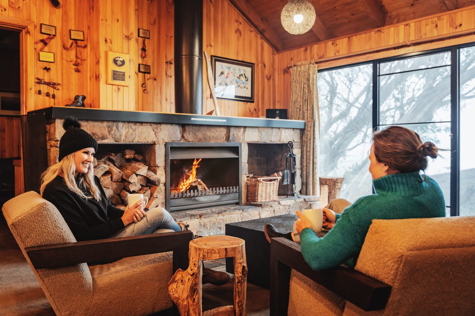 Two women sitting in lounge chairs by a log fire in a wooden ski club, drinking coffee from white mugs and smiling as they chat.